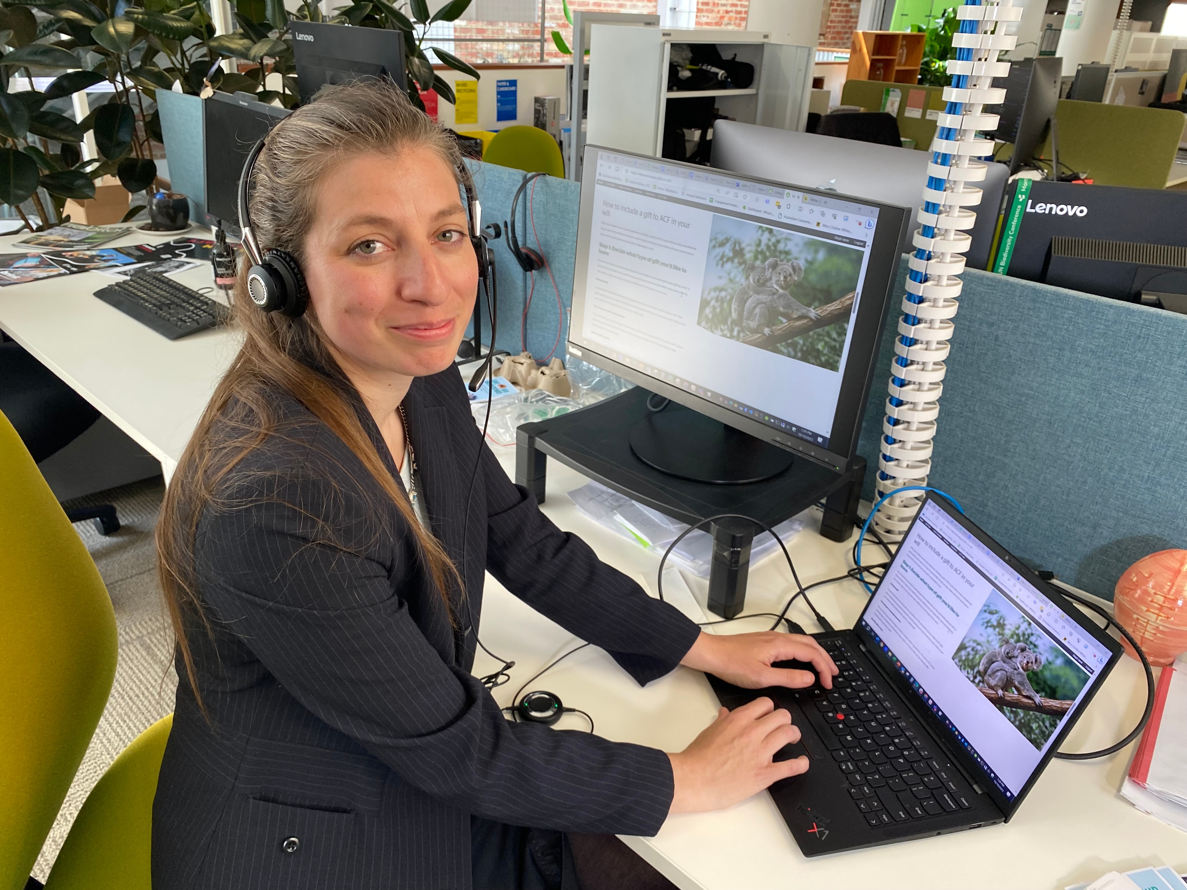 A woman with long brown hair and wearing a headset sits in front of a laptop and desktop computer.