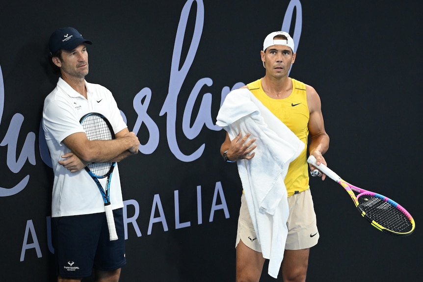 Carlos Moya stands against a wall with a racket to his chest as Rafael Nadal towels himself at the Brisbane International