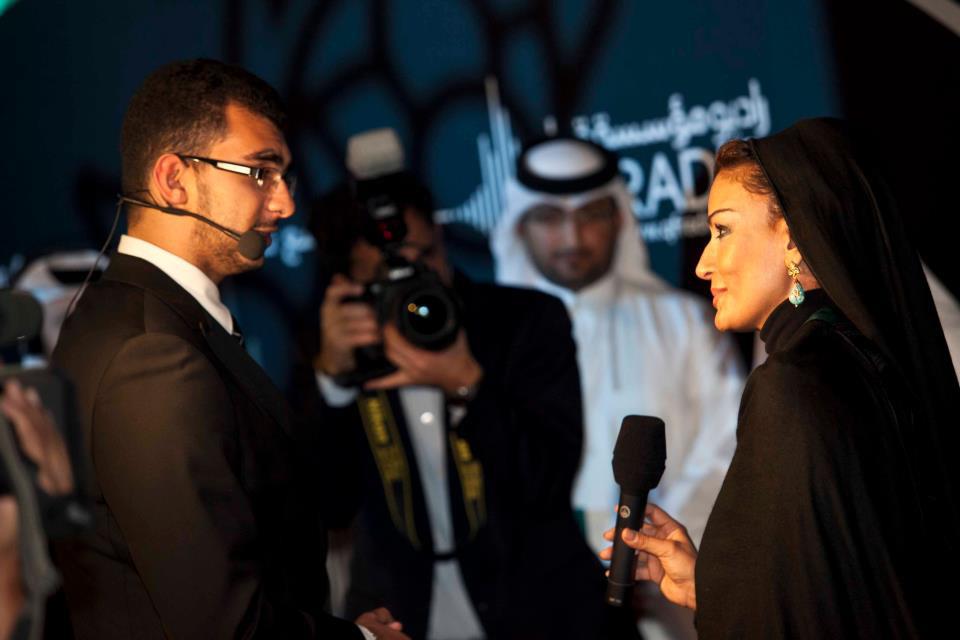 Man with microphone headset interviewing a woman in black headscarf and dress as a photographer takes a photo.
