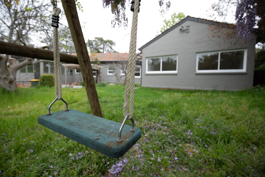 An empty swing, outside a grey-coloured home.
