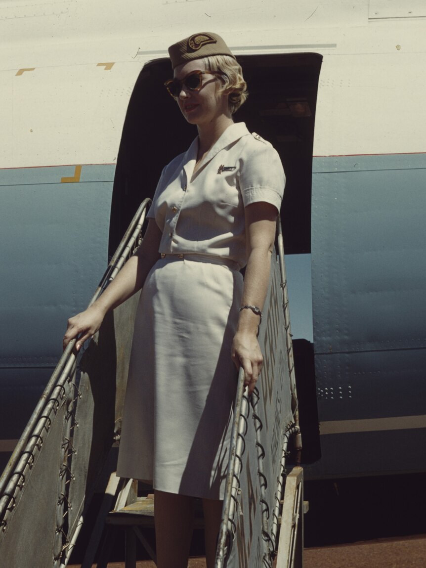 An air hostess in a pale dress stands on the steps to an airplane in 1962.