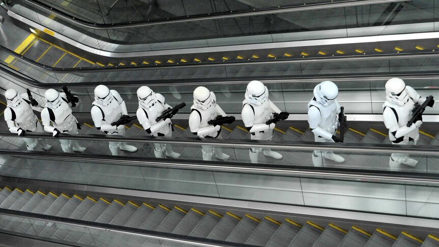 A line of Imperial Stormtroopers, from the Star Wars movies, travel up an escalator.