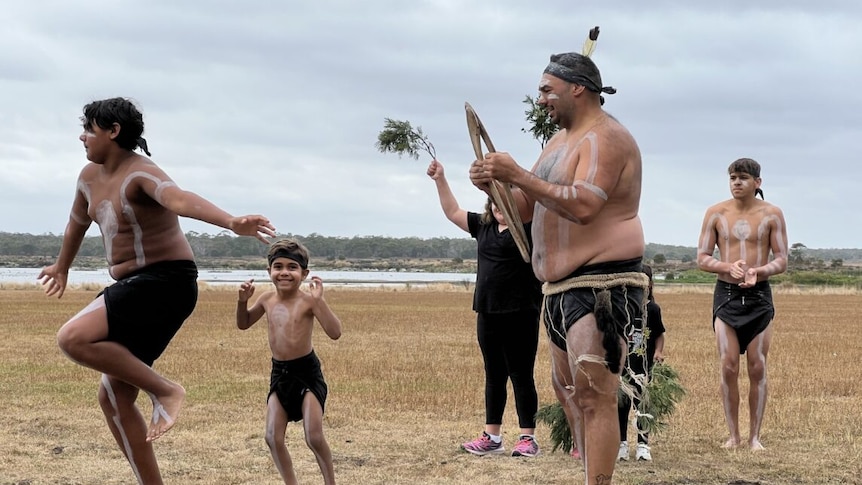 Indigenous men and children in traditional garb dancing near the ocean.