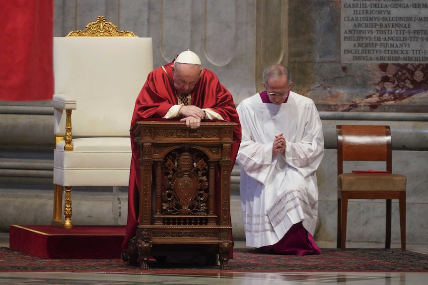 The Pope, in a red gown and white cap, kneels behind a wooden lectern, head bowed in prayer.