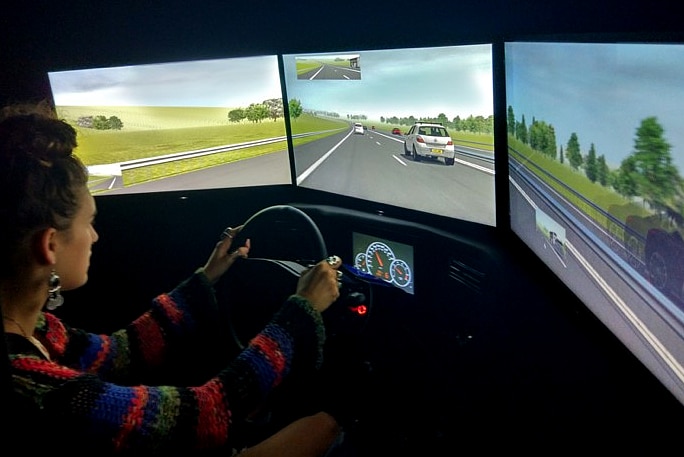 A woman sits at a driving simulator looking at screens showing animated version of cars on a road.