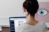A woman sits at a table typing on a laptop in front of a white wall. An artwork next to her of an eyeball appears to look at her