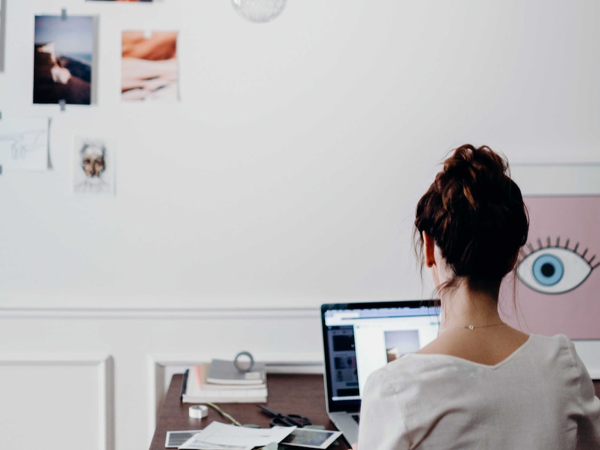 A woman sits at a table typing on a laptop in front of a white wall. An artwork next to her of an eyeball appears to look at her