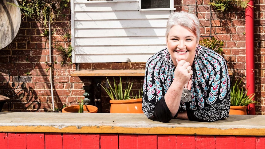 Kelly Higgins-Devine, smiling, standing at the back of a brick and weatherboard house.