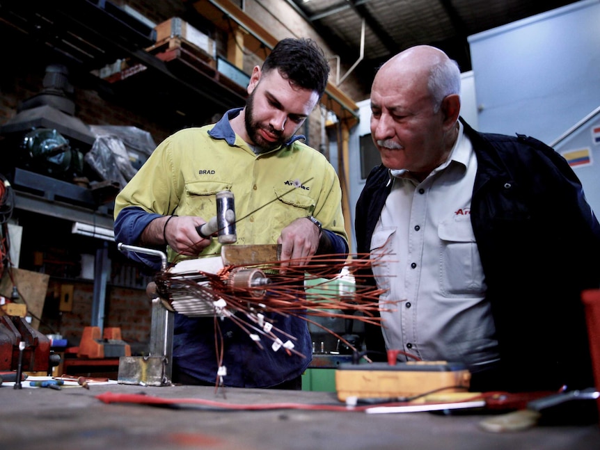 A young man wearing yellow high vis works on some copper wiring while an older man with a moustache watches next to him