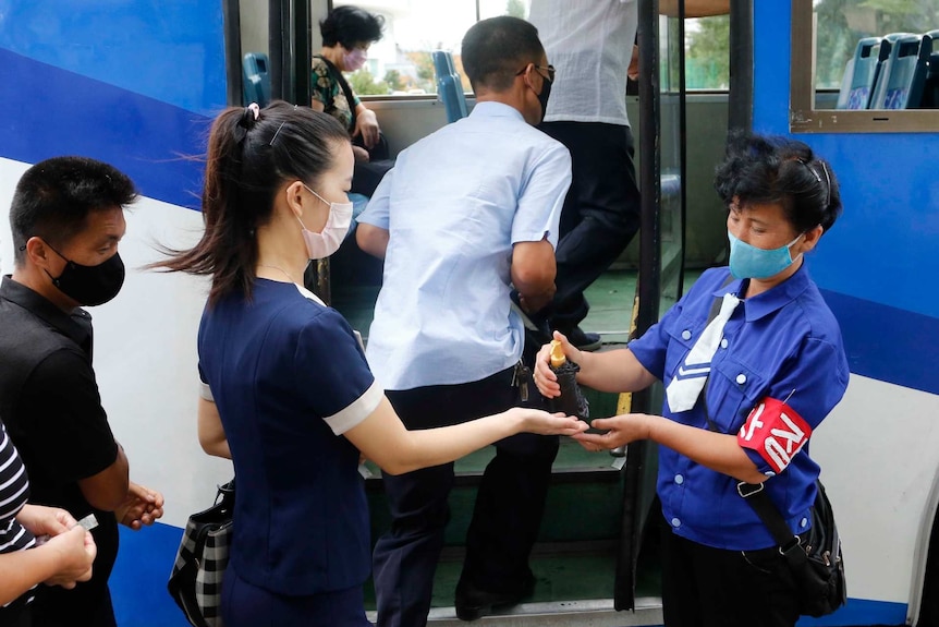 A passenger wearing a face mask has her hands disinfected by a person in a tie as she waits to board a bus
