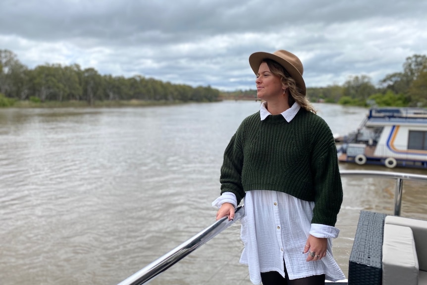 A woman wearing a white shirt, brown jumper and wide-brimmed hat, holding a rail looking out to the river