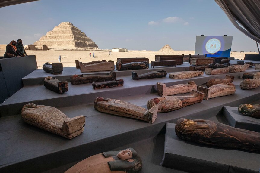 a very large number of sarcophagi laide out on the floor. In the far background is an Egyptian pyramid.