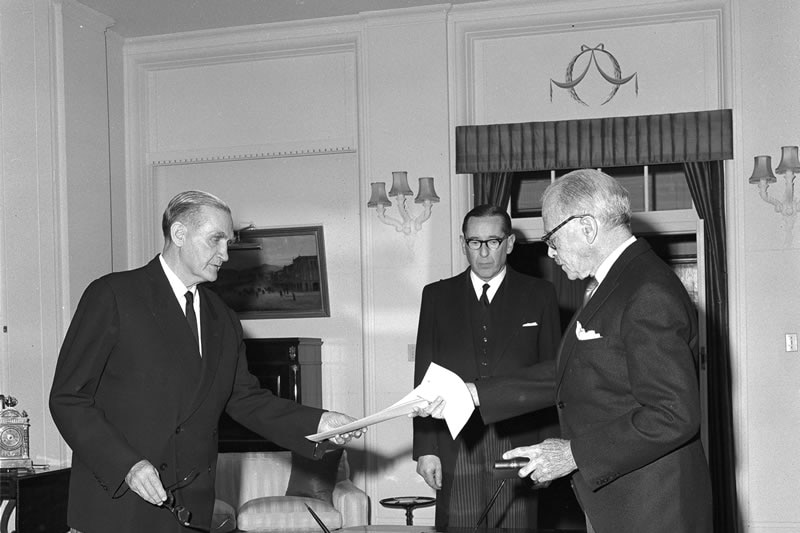 Three men in suits, one hands a document to the other, black and white image