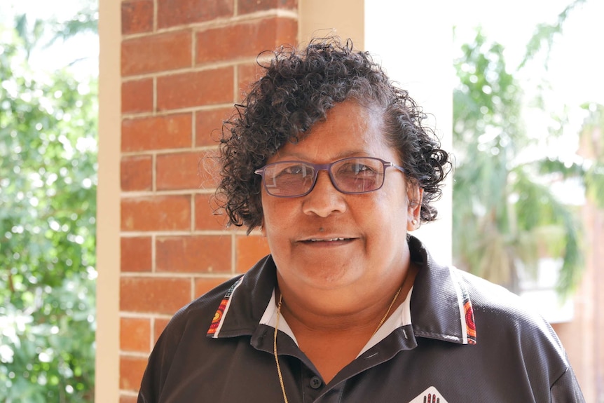 A woman wearing reading glasses and a gold chain smiles at the camera. In the background is a red brick wall and palm trees.