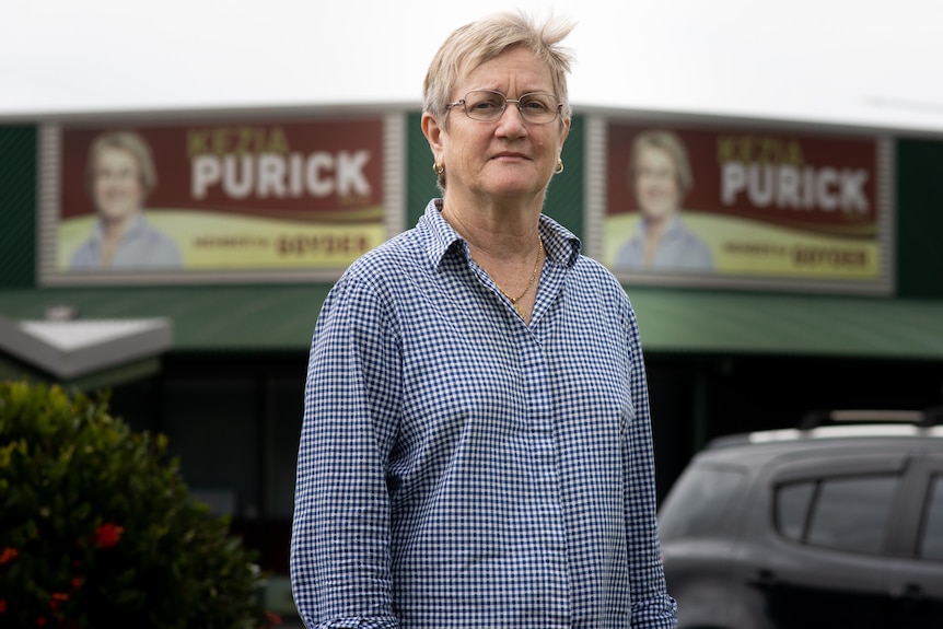 A woman in a pattered button-down shirt looks seriously at the camera. She has short, blonde hair.