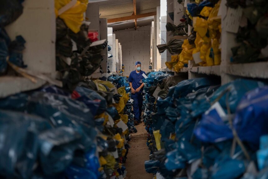 a man with a face mask on stands inside an ossuary filled with blue and yellow bags