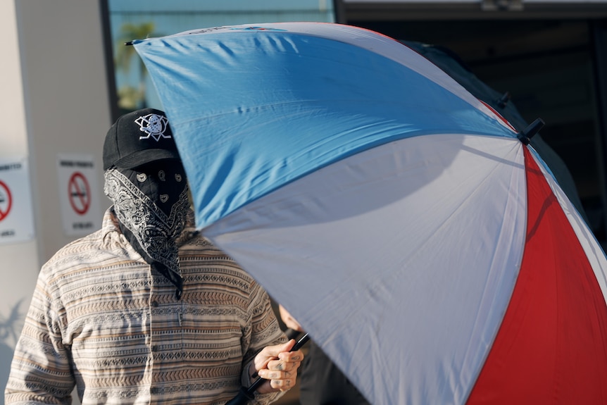 Man with bandana covering face holds up an umbrella outside court 