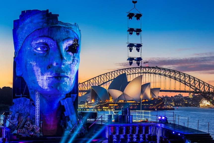A giant blue head and bust of an Egyptian god looms over a stage set on Sydney harbour with the Opera House and bridge behind.
