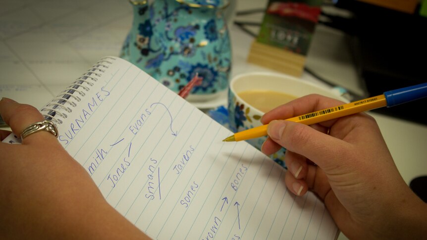 Hands hold a notebook with surnames written on it, a cup of tea in the background.