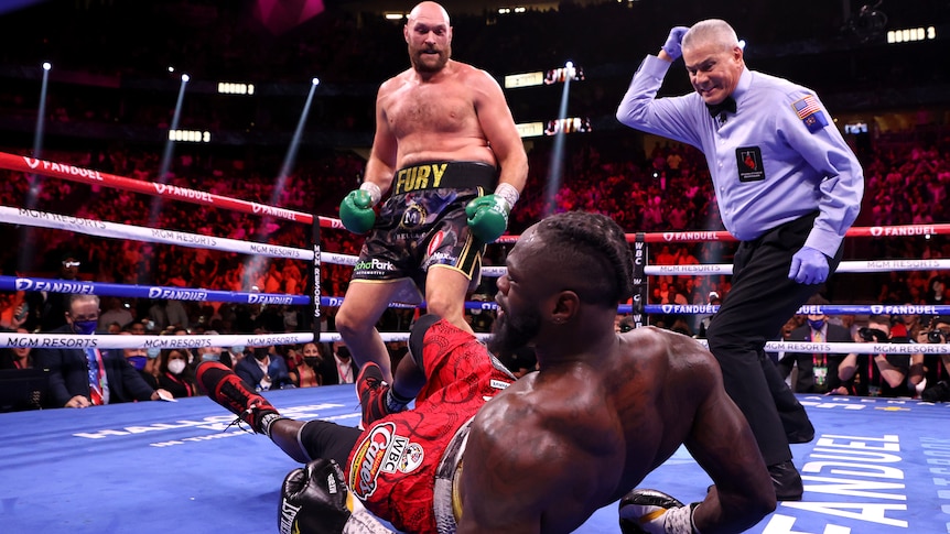 Tyson Fury looks down at Deontay Wilder who has fallen to his back. The referee watches on