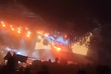 A slightly blurry screenshot shows a metal stage structure leaning at an awkward angle over a crowd, mid-collapse.