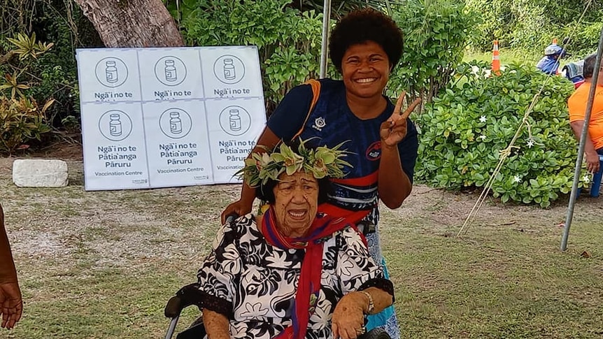 A woman smiles and holds up peace sign while standing behind older woman in wheelchair outside but under cover.