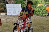 A woman smiles and holds up peace sign while standing behind older woman in wheelchair outside but under cover.