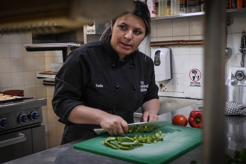 Une femme sérieuse en uniforme de chef noir coupant du capsicum dans une cuisine commerciale, regarde loin de la caméra.