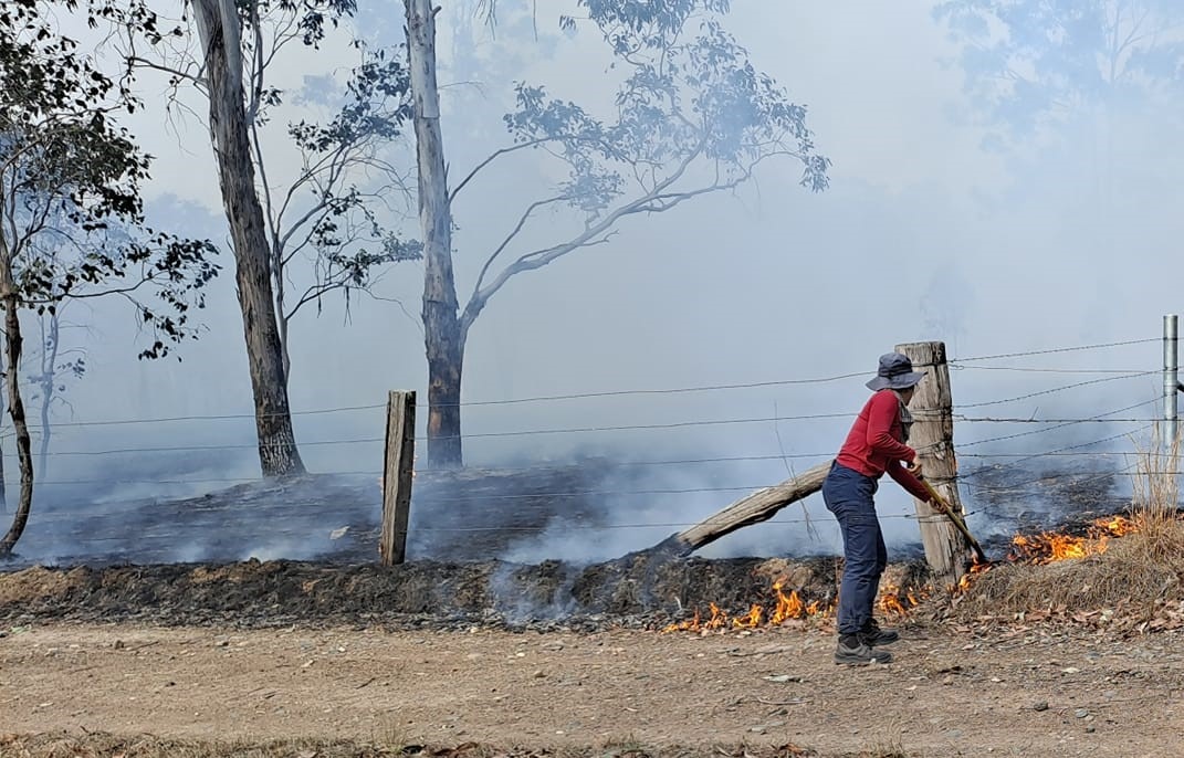A woman shoveling dirt onto a burning fire near the fence line, smoke obscuring the horizon. 