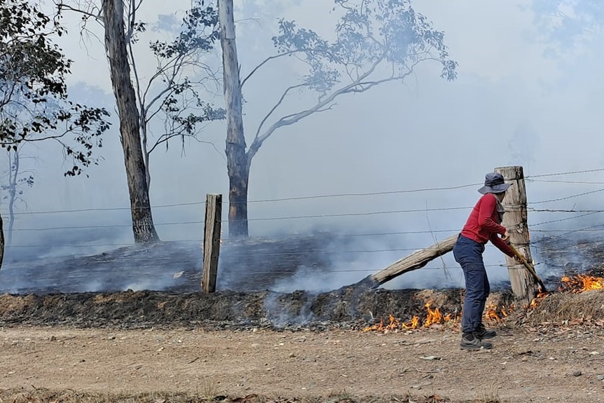A woman shoveling dirt onto a burning fire near the fence line, smoke obscuring the horizon. 