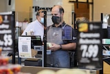 A man working at a supermarket checkout wearing a black mask.