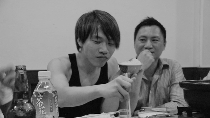 一名年轻男孩坐在桌边吃火锅，一名年长男子在他身后看着他