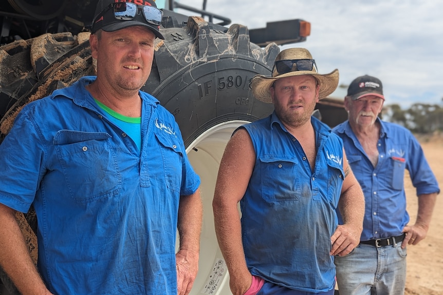 Three fair-skinned male farmers in blue work shirts stand next to large machinery 