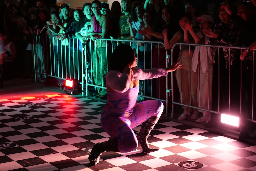 A dancer squats in heels in front of a red light, to pose in front of a crowd. 