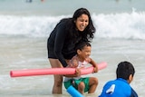 A woman laughs and plays with her young sons in the shallow waves at the beach.