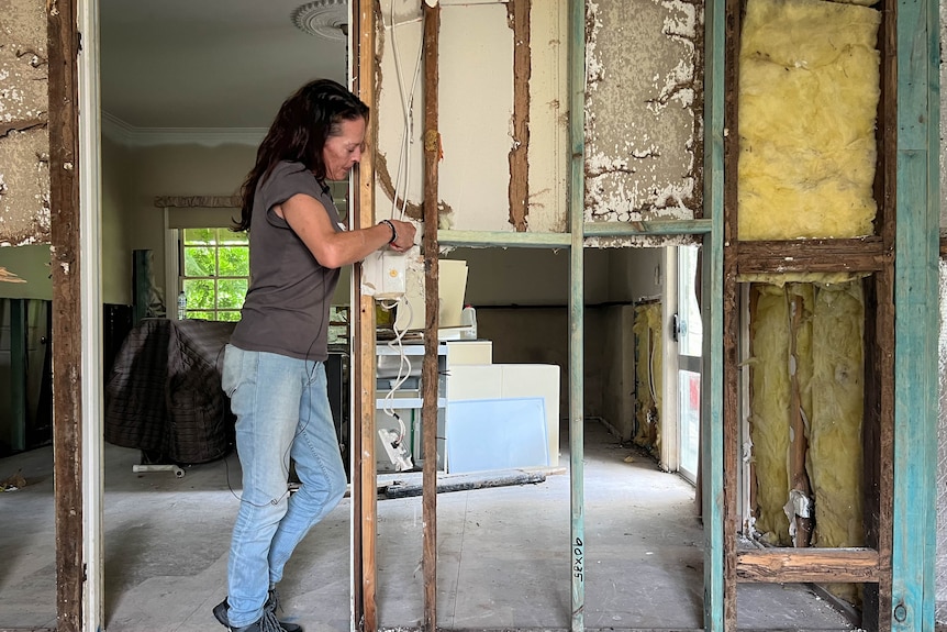 A woman stands in a doorway cutting plasterboard, revealing the rest of the house behind the studs in the wall