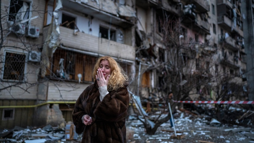 A woman in winter clothing cries while standing outside of a badly damaged block of apartments.