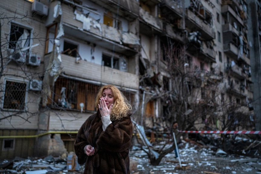 A woman in winter clothing cries while standing outside of a badly damaged block of apartments.