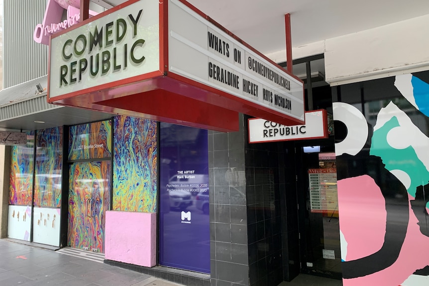 Un grand panneau électronique indiquant Comedy Republic est suspendu au plafond devant un mur coloré.