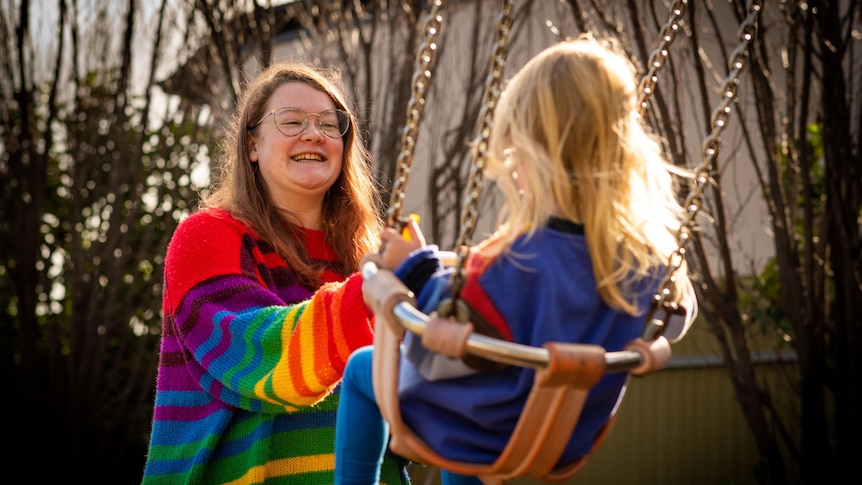 A woman in a bright colourful jumper pushes her young child on a swing.