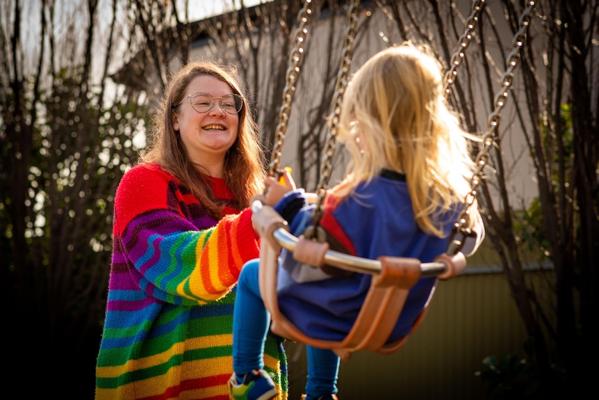 A woman in a bright colourful jumper pushes her young child on a swing.