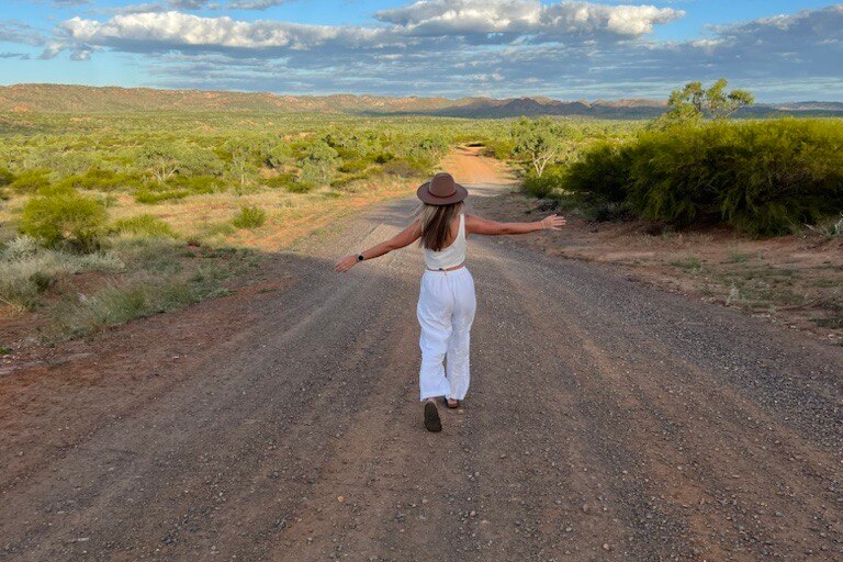 A young blonde woman walks down an outback road with her arms outstretched.