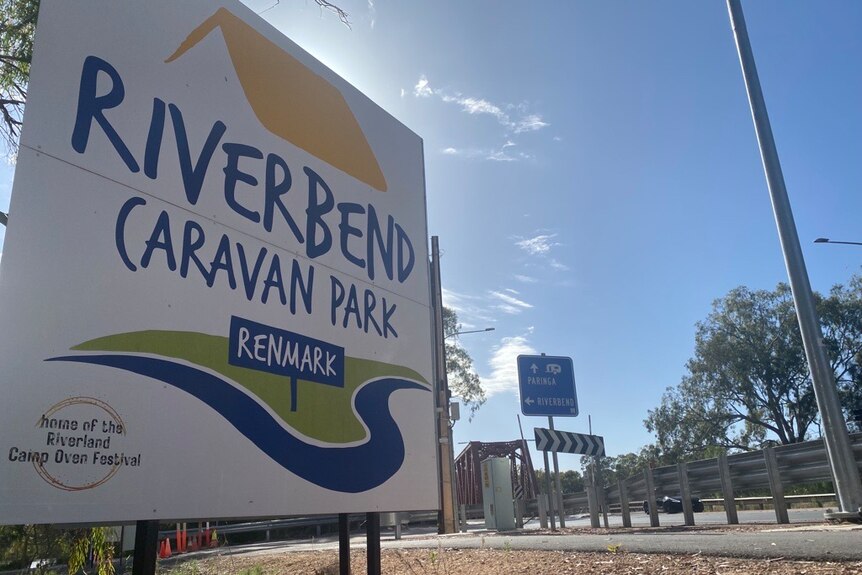 A picture of a sign of a caravan park in the riverland with a road behind it and other signage. 