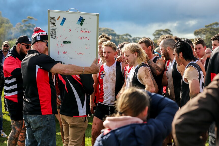 A group of football players wearing red white and black stand on ground looking at whiteboard.