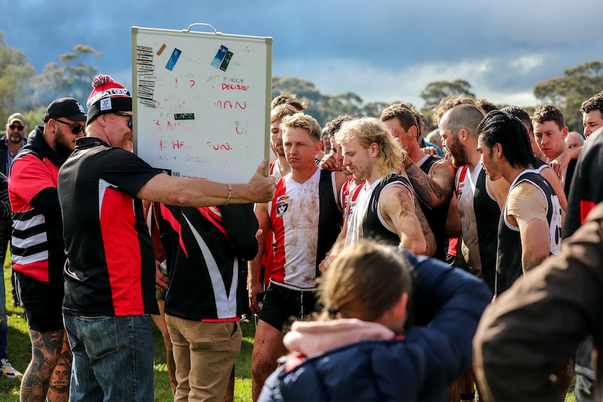 A group of football players wearing red white and black stand on ground looking at whiteboard.