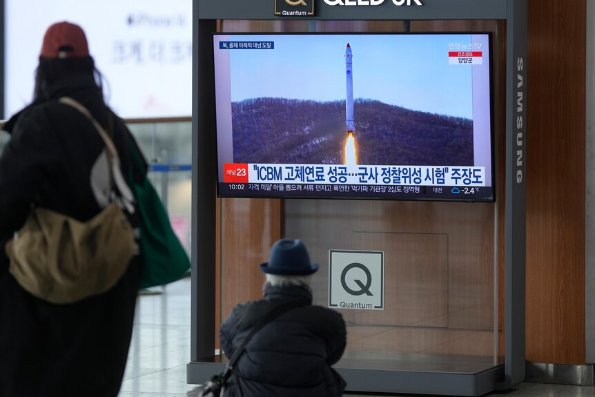 两个亚洲人在看电视，屏幕上是一枚尾部喷着火焰的导弹。