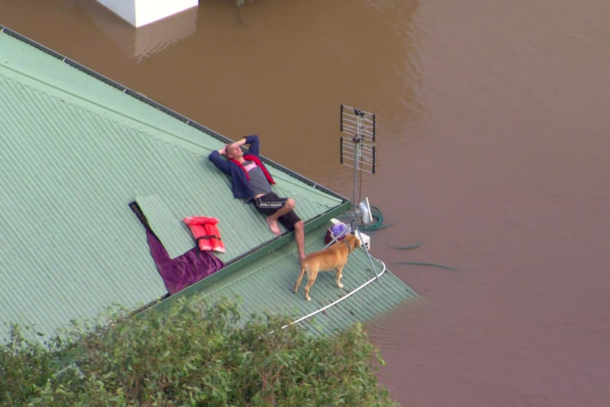 A man sits on the roof of his house with a pet dog surrounded by floodwaters.