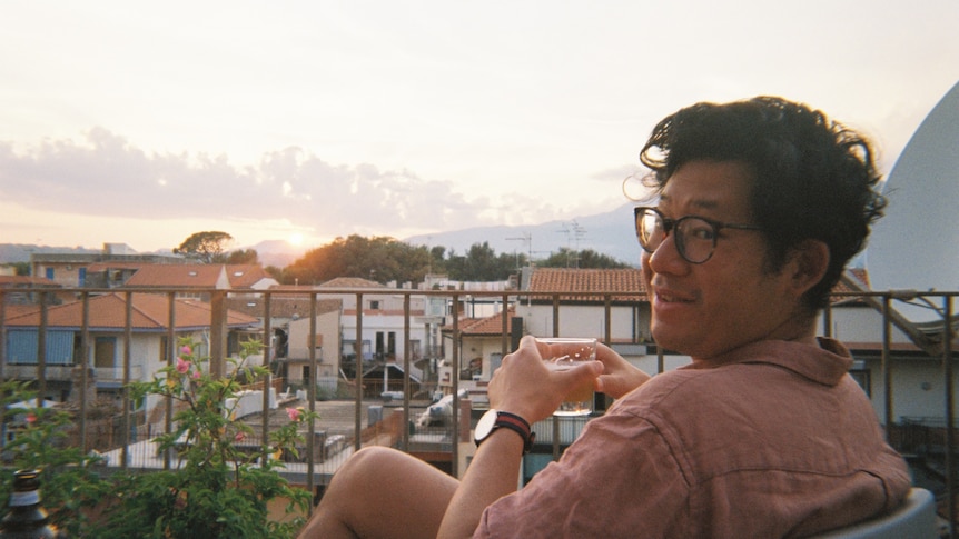 Author André Dao sitting on a balcony at the end of the day, smiling as he holds a drink. He wears a linen shirt and glasses.