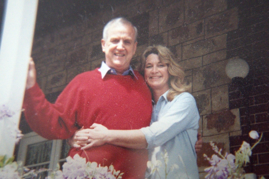 A man in a red jumper is hugged by a woman in a blue shirt, both smiling at the camera.