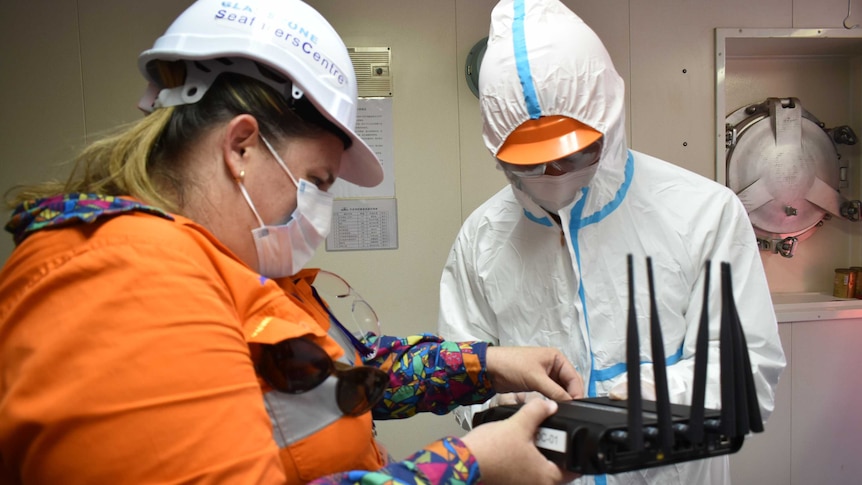 Woman in orange shirt, wearing mask, hard hat, another unidentified person in full PPE both setting up Wi-Fi modem.